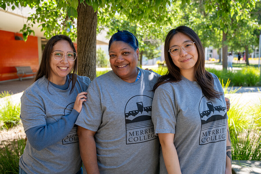 Three smiling individuals standing outdoors under a tree, wearing matching gray t-shirts with the Merritt College logo.